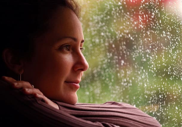 2 Small Ways to Start Living the Life You Truly Want Woman looking out of rainy window with a slight smile on her face.