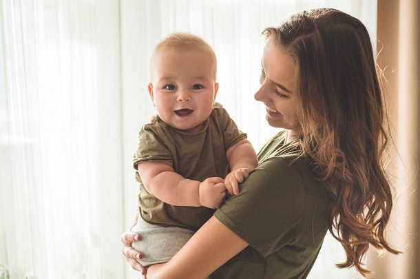 97 Funny Mother’s Day Quotes for Laughter, Relatable Humor and Gratitude A smiling mother holding her baby who looks into the camera with a smile.
